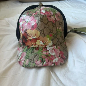 Gucci blommig keps med mesh - Snygg keps från Gucci med klassiskt GG-mönster och färgglada blommor i rosa, vitt och orange. Framsidan är i canvas med blommigt tryck, baksidan har svart mesh och justerbar plastrem. Perfekt statement-piece för dig som vill sticka ut.