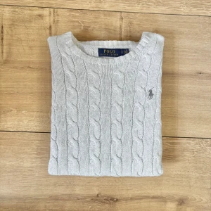 Ljusgrå kabelstickad tröja Polo Ralph Lauren - Kabelstickad tröja från Polo Ralph Lauren i ljusgrått med klassisk rund halsringning och broderad logga på bröstet. Tröjan är i mjuk bomull, har ribbade muddar och lång ärm. Perfekt för lager-på-lager och snygg till jeans.