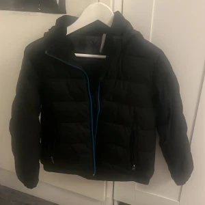Svart pufferjacka från Everest - Svart pufferjacka från Everest med huva och blå dragkedja. Jackan har quiltad design, två fickor med dragkedja och är perfekt för kalla dagar. Snygg och enkel stil som funkar till det mesta.