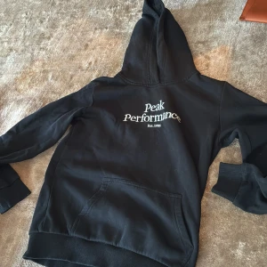Svart hoodie från Peak Performance - Svart hoodie från Peak Performance med vit logga på bröstet. Klassisk känguruficka framtill, ribbade muddar och stor huva. Perfekt för en avslappnad och sportig stil.