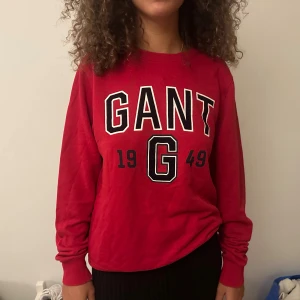 Röd sweatshirt från GANT med tryck - Röd sweatshirt från GANT med stort svartvitt tryck framtill med texten 'GANT 1949'. Tröjan har rund halsringning, långa ärmar och ribbade muddar vid ärmslut och nederkant. Perfekt för en chill och sportig look.