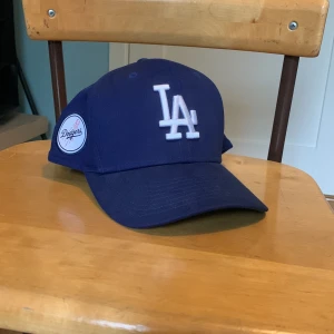 Blå LA Dodgers keps - Snygg blå keps från LA Dodgers med vit broderad LA-logga framtill och Dodgers-märke på sidan. Kepsen har böjd skärm med grön undersida och justerbar snapback baktill. Skriv vid minsta lilla fundering!