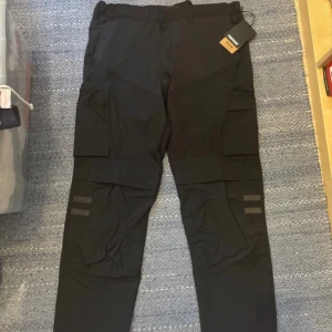 Svarta cargopants från Fristads C54 - Svarta cargopants från Fristads i storlek C54. Byxorna är nya aldrig använda med lapparna kvar. Nya priset på byxorna är 1299 kr. Byxorna har flera praktiska fickor på sidorna, stretchigt material och diskreta reflexdetaljer på benen. Perfekta för dig som gillar funktionella och snygga byxor med modern look. 