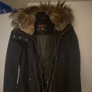 Svart parkas från Woolrich med päls - Säljer en svart parkas från Woolrich med stor fluffig päls runt huvan och snygga guldfärgade dragkedjor. Jackan har en klassisk lång modell och är perfekt för kalla dagar. Fodret är mjukt och jackan har flera praktiska fickor och detaljer.