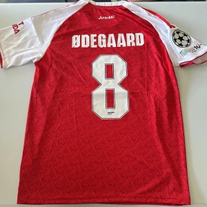 Arsenal Ødegaard 8 matchtröja Adidas - Säljer en officiell Arsenal fotbollströja med Ødegaard och nummer 8 på ryggen. Tröjan är röd med vita ärmar, har Champions League-märke på ärmen och tydligt Arsenal-emblem framtill. Tillverkad i lätt funktionsmaterial med diskret mönster och klassiska Adidas-ränder.