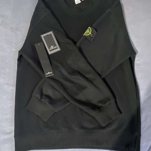 Svart sweatshirt från Stone Island - Svart sweatshirt från Stone Island med klassisk rund hals och ribbade muddar. Tröjan har den ikoniska Stone Island-loggan på vänster ärm. Tillverkad i mjuk bomull, perfekt för en clean och stilren look.