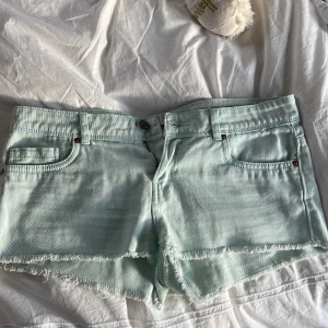 Ljusblå jeansshorts med fransig kant - Säljer ett par ljusblå jeansshorts med rå, fransig kant nertill. Klassisk femficksmodell med bälteshällor och knappstängning. Perfekta för varma dagar och ger en avslappnad vibe.