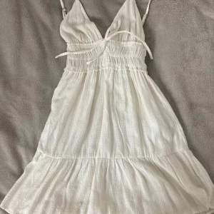 Vit kort sommarklänning Pull&Bear XS - Supersöt vit kort klänning från Pull&Bear i storlek XS. Klänningen har smala axelband, omlott-v-ringning och knyt i midjan. Lätt rynkad midja och volang nedtill ger en luftig känsla. Perfekt för varma dagar och har en diskret struktur i tyget.