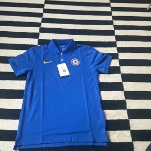 Chelsea Nike blå Piké S. - Chelsea FC piké från Nike i blått, tillverkad i Dri-FIT-material för bästa komfort. Slim fit-modell med klassisk krage och korta ärmar. Broderad Chelsea-logga på bröstet och gul Nike Swoosh. Perfekt för dig som vill visa ditt stöd för laget.  Helt äkta, aldrig använd. Helt ny.
