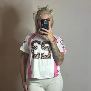 Vit t-shirt med leopardtryck och rosa detaljer - Vit t-shirt med stora leopardmönstrade siffror 23 fram och bak, rosa ränder längs sidorna och stjärntryck på ärmarna. Modellen är kortärmad och har en loose fit. Perfekt för dig som gillar sportig och edgy stil.