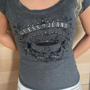 Grå t-shirt från Guess Jeans - Snygg grå t-shirt från Guess Jeans med tryck och pärldetaljer framtill. Modellen är figurnära med korta ärmar och rund halsringning. Perfekt för dig som gillar en stilren men ändå cool look. 2000s stil säljs för 250 storlek xs