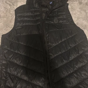 Svart dunväst från Jack & Jones - Snygg svart dunväst från Jack & Jones med quiltad design och dragkedja framtill. Västen har en stilren look och passar perfekt över en hoodie eller tröja när du vill hålla värmen utan att tumma på stilen.