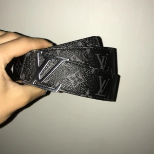  svart monogram bälte - Säljer ett bälte i svart med klassiskt monogrammönster och silverfärgat spänne. Bältet är gjort i skinn med beige insida och tydliga logodetaljer. Perfekt accessoar för att lyfta din outfit med en lyxig touch.