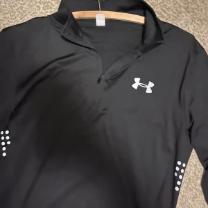  långärmad träningströja Under armour - Svart långärmad träningströja från Under Armour med halv dragkedja och reflekterande detaljer på ärmarna. Perfekt för löpning eller gym. Tillverkad i lätt och stretchig polyester som andas. UA-logga på bröstet och hög krage för extra komfort.