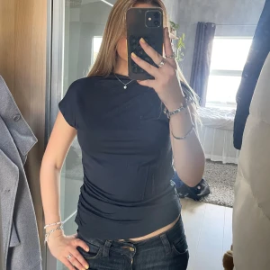 Mörkblå tight t-shirt med kort ärm - Enkel och stilren svart t-shirt med tight passform och korta ärmar. T-shirten har en rak halsringning med ”scrunch” på sidan. Är gjord i ett mjukt material som sitter snyggt mot kroppen. Perfekt till jeans eller kjol för en clean look.