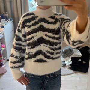 Zebramönstrad fluffig polotröja - Supermjuk och fluffig polotröja med svartvitt zebramönster. Tröjan har hög krage, breda ärmar och är stickad i ett gosigt material som känns riktigt cozy. Perfekt för kyliga dagar när du vill sticka ut lite extra.