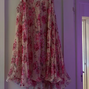 Blommig kortklänning från Dainty - Superfin kortklänning från Dainty i ett luftigt, transparent material med volangig nederkant. Klänningen har ett romantiskt blommönster i rosa och lila toner på en ljus bas. Perfekt för sommarens alla tillfällen när du vill känna dig extra söt.