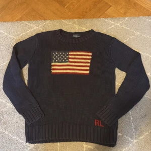 Mörkblå stickad tröja Polo Ralph Lauren flagga - Mörkblå stickad tröja från Polo Ralph Lauren med amerikansk flagga på bröstet och RL-broderi i rött vid nederkanten. Klassisk rund halsringning och långa ärmar. Perfekt för dig som gillar preppy stil och vill sticka ut med en ikonisk detalj. Det är storlek L i barn så motsvarar typ xs i vuxen.