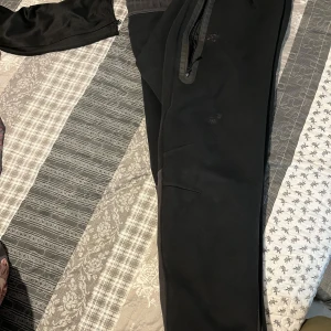 Svarta mjukisbyxor från Nike - Svarta mjukisbyxor från Nike med klassisk logga på benet. Byxorna har dragkedjeförsedda fickor och ribbade muddar vid bensluten. Tillverkade i mjukt material som är perfekt för chill eller träning. Snygga detaljer med mörkare paneler längs sidorna.