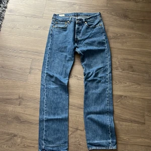 Levi's 501 blå jeans W31 L34 - Klassiska Levi's 501 jeans i blå denim med raka ben och normal passform. Byxorna har fem fickor, knappgylf och den ikoniska läderpatchen bak i midjan. Snygga kontrastsömmar och röd Levi's-tagg på bakfickan. Perfekta för en avslappnad stil.