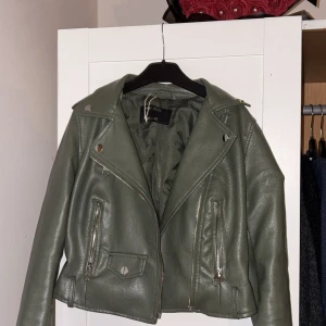 Olivgrön skinnjacka från Sinsay XXS - Snygg olivgrön skinnjacka från Sinsay med klassisk bikerstil, dragkedjor och silvriga knappar. Jackan har långa ärmar, bred krage och coola detaljer som fickor med dragkedja och knapp. Perfekt för dig som vill ha en edgy look.