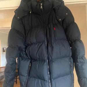 Marinblå dunjacka från Polo Ralph Lauren - Säljer en marinblå dunjacka från Polo Ralph Lauren med hög krage, avtagbar huva och dragkedjor på fickorna. Jackan har en klassisk pufferstil och den ikoniska röda loggan på bröstet. Perfekt för kalla dagar när du vill hålla stilen.