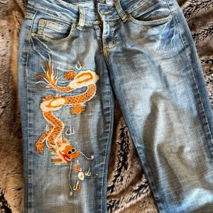 Energie Young jeans med drakbrodyr - Blå jeans från Energie Young med broderad drake på ena benet och broderade palmer samt text på bakfickorna.🌴🐉 Klassisk femficksmodell med raka ben och snygga slitningar på byxorna. Passar mig som är 170 cm lång. Fållorna längst ned på bena är utvikta och ned strykna för att de ska vara längre i benen längden. Unika och häftiga jeans🙌