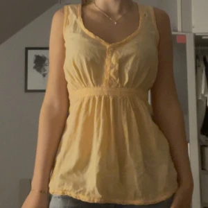 Blus  - Säljer en söt och luftig blus i ljus persikoton med v-ringning och knappar framtill. Toppen är ärmlös och har lätt rynk vid midjan för en avslappnad passform. Perfekt för varma dagar och enkel att matcha med jeans eller kjol.