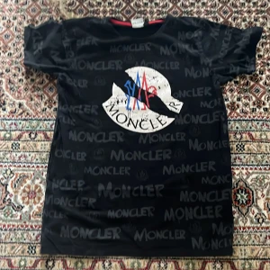 Svart Moncler t-shirt med logga - Svart t-shirt från Moncler med stor vit logga framtill och tryckt Moncler-text över hela plagget. Klassisk rund hals med röd kant och korta ärmar. Snygg streetstil med inslag av blått och rött i loggan. Perfekt för dig som gillar exklusiva märken och vill sticka ut.