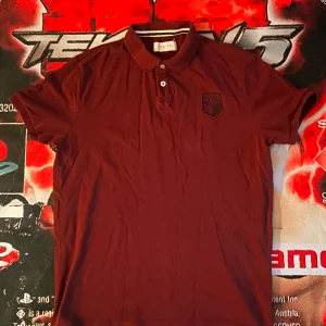 Vinröd pikétröja från Pier One - Vinröd pikétröja från Pier One med klassisk krage och två knappar framtill. Tröjan har korta ärmar och en broderad logga på bröstet. Perfekt för en chill och stilren look.