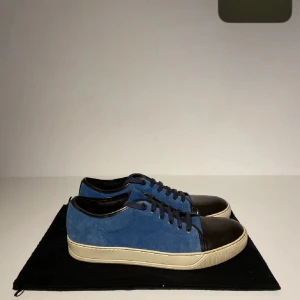 Blå Lanvins - Snygga sneakers från Lanvin med blå mocka på sidorna och svart skinn på tån och runt öppningen. Skorna har svarta snören och en beige platt sula. Perfekt för dig som gillar stilrena och exklusiva sneakers med en unik färgkombination.