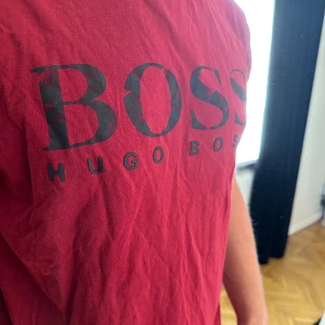 Röd Hugo Boss t-shirt med tryck - Snygg röd t-shirt från Hugo Boss med stort svart BOSS-tryck på bröstet och mindre logga i nacken. Klassisk rund hals och korta ärmar. Mjuk bomullskvalitet som är skön mot huden. Perfekt för dig som gillar stilrena märkesplagg.