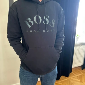 Svart hoodie från Hugo Boss - Svart hoodie från Hugo Boss med stor logga framtill. Tröjan har huva med snörning och en stor magficka. Tillverkad i mjuk bomull och har långa ärmar med ribbade muddar. Perfekt för en chill och stilren look.