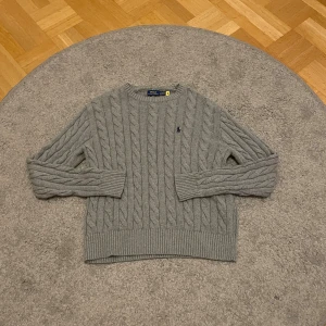 Grå kabelstickad tröja Polo Ralph Lauren - Kabelstickad grå tröja från Polo Ralph Lauren med klassisk rund hals och broderad marinblå logga på bröstet. Inga defekter alls på tröjan och priset är INTE hugget i sten vid snabb affär. Skriv vid minsta fundering! 