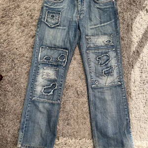 Blå patchade jeans från Daniel Jeanswear - Unika jeans från Daniel Jeanswear med patchade detaljer och slitningar. Ger en riktigt edgy look.Slitna jeans med vida ben. Baggy jeans. Model 1015. W: 32, L:34 100% bomull Det är nog en herrmodell, men passar mig som kvinna bra 