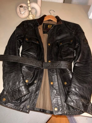 Brun skinnjacka Belstaff Gold Label - Brun skinnjacka från Belstaff Gold Label med bälte i midjan, fyra stora fickor med knappar och Manchester detaljer vid kragen. Jackan har en klassisk look med guldfärgade knappar och beige innerfoder. Perfekt för dig som gillar tidlös stil och coola detaljer.