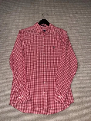 Rutig röd skjorta från GANT - Säljer en snygg röd och vit rutig skjorta från GANT i storlek M. Skjortan har klassisk krage, vita knappar och ett broderat GANT-logo på bröstfickan. Regular fit och tillverkad i mjuk bomull. Perfekt för dig som gillar stilrena och tidlösa plagg.