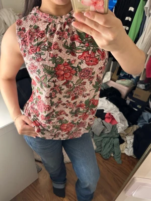 Blommig ärmlös blus i rosa och rött - Säljer en söt blommig blus med ärmlös design och rundad hals. Toppen har ett mönster med röda och rosa blommor samt gröna blad på en ljusrosa bakgrund. .