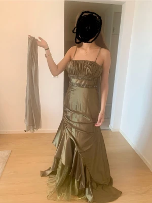 Olivgrön långklänning med pärldetaljer - Säljer en olivgrön långklänning med smala axelband och vackra pärldetaljer runt midjan. Klänningen har ett glansigt tyg med draperad överdel och volang i nederdelen. Materialet känns lyxigt och skimrande, perfekt för bal eller fest. Medföljer matchande sjal. Köpte för 2400kr. Använd 1 gång.
