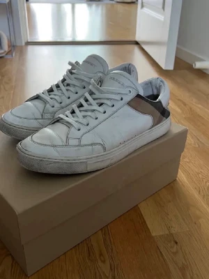 Burberry vita sneakers med rutigt tyg - Säljer ett par snygga vita sneakers från Burberry med klassiskt rutigt tyg på hälen och sidan. Skorna har snörning, rund tå och platt sula. Materialet är skinn och textil, och de kommer med originalkartong och dustbag. Perfekta för dig som gillar stilrena designer.