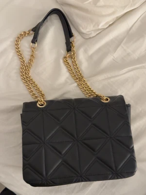 Svart quiltad axelväska med guldkedja - Snygg svart axelväska med quiltat geometriskt mönster och guldfärgade kedjor som axelrem. Väskan har en stilren och modern look. Aldrig använd 