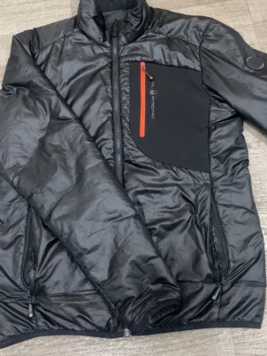 Svart pufferjacka från Sail Racing - Svart pufferjacka från Sail Racing med orange dragkedja på bröstfickan och snygga detaljer. Jackan har hög krage, flera dragkedjeförsedda fickor och en stilren badge på ärmen. Materialet är syntet och polyester, perfekt för kyliga dagar. OBS! Finns ett litet hål vid ena armen, men den är fint fixad så det är inget man märker.