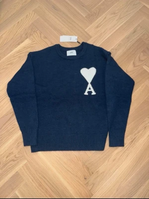 Mörkblå stickad tröja från Ami Paris  - Snygg mörkblå stickad tröja från Ami med ett stort vitt hjärta och bokstaven A på bröstet. Tröjan har rund hals, ribbade muddar och lång ärm. Perfekt för dig som gillar stilrena detaljer och vill ha något som sticker ut lite. 