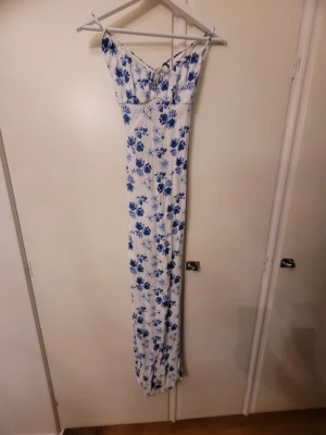 Vit långklänning med blå blommor FB Sister - Supersnygg långklänning från FB Sister i vitt med blått blommigt mönster. Klänningen har smala axelband, öppen rygg med snörning och omlottdetalj. Perfekt för dig som gillar en feminin och fräsch stil. Materialet känns lätt och luftigt.