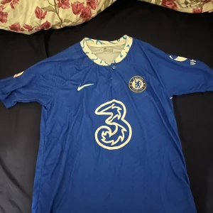 Chelsea FC Nike matchtröja blå - Chelsea FCs officiella matchtröja från Nike i klassisk blå färg med klubbmärke på bröstet och stor sponsorlogga framtill. Tröjan har korta ärmar och mönstrad krage med vita och blå detaljer. Perfekt för dig som älskar fotboll och vill visa ditt stöd för laget.
