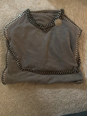Grå axelväska från Stella McCartney - Säljer en snygg grå axelväska från Stella McCartney med kedjedetaljer runt kanterna och som axelrem. Väskan är tillverkad i mjuk mocka och har en rund medaljong med logga. Perfekt storlek för allt du behöver. Åkthetsbecis går att leta fram. pris kan diskuteras. Skriv för fler frågor🩷