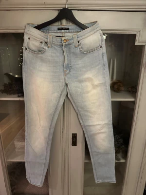 Ljusblå jeans från Nudie Jeans - Säljer ett par ljusblå jeans från Nudie Jeans med klassisk femficksdesign och snygga slitningar. Jeansen har raka ben och orangea sömmar som ger en schysst kontrast. Passar dig som gillar en clean och avslappnad stil.