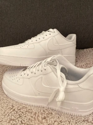 Nike Air Force 1 helvita sneakers - Nike Air Force 1 sneakers i helvit färg med klassisk låg siluett. Skorna har snörning, perforerad tå och rund tå. Tillverkade i skinn med platt sula och ikonisk Nike-logga på sidan. Perfekta för dig som gillar cleana och stilrena sneakers.