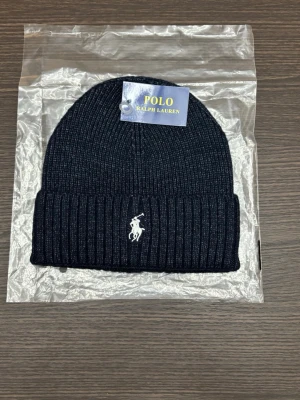 Svart ribbad mössa från Polo Ralph Lauren - Snygg svart ribbstickad mössa från Polo Ralph Lauren med klassisk broderad logga i vitt framtill. Mössan har uppvikt kant och är tillverkad i mjukt stickat material som håller dig varm och stilren under kyliga dagar.