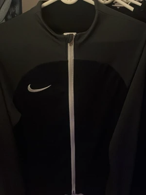 Svart Nike Dri-Fit zip-tröja slim fit - Nike Dri-Fit zip-tröja i svart med vit dragkedja och vit Nike-logga på bröstet. Slim fit-modell med sportig look, tillverkad i syntetmaterial som andas. Perfekt för träning eller chill. Snygg och stilren design med lång ärm.